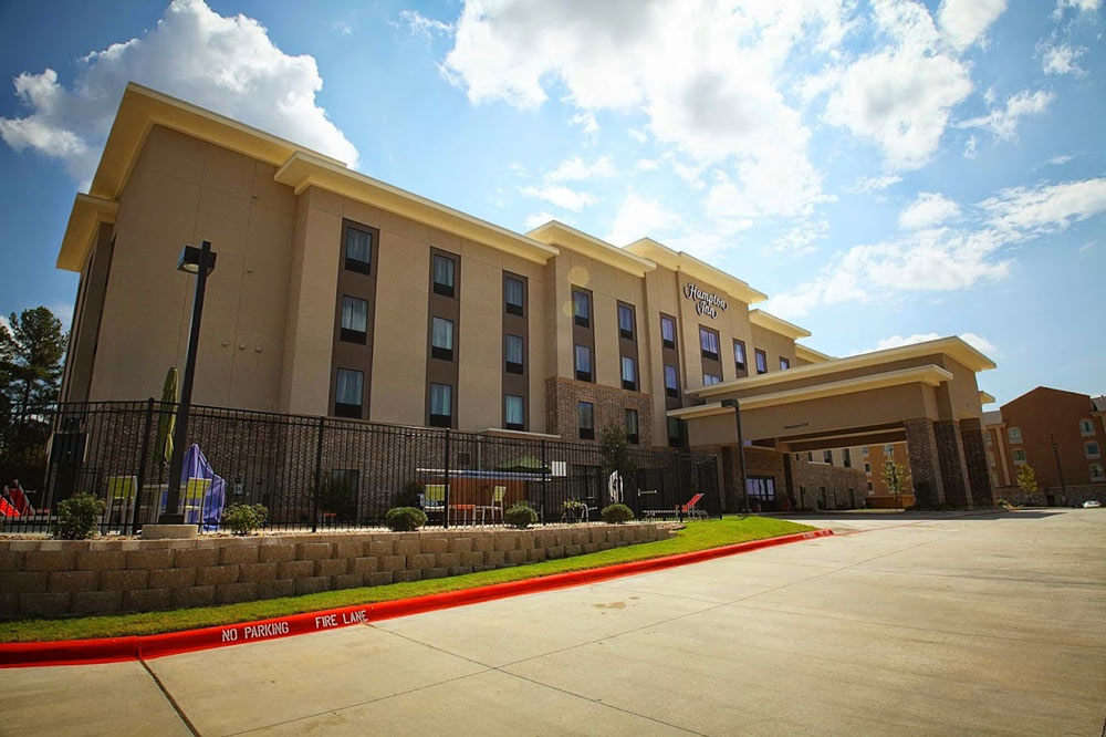 Exterior of Hampton Inn in Texarkana, showcasing a modern, multi-story building with a covered entrance, parking area, and sunny sky in the background.
