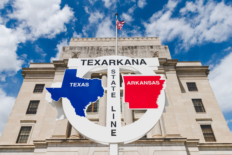 Texarkana State Line marker with Texas and Arkansas outlines in front of the historic U.S. Post Office building, under a blue sky with clouds.