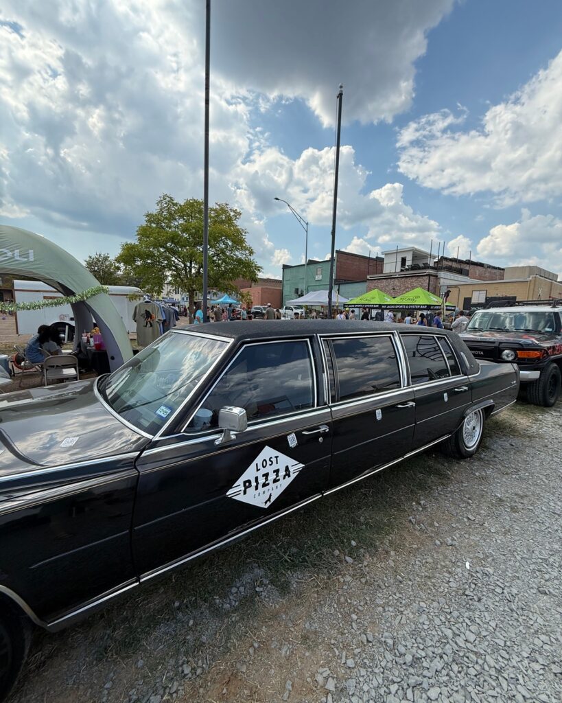 Black Lost Pizza Company car parked at Texarkana Pickle Fest 2025 with vendor tents and festival crowds in downtown Texarkana, Arkansas.