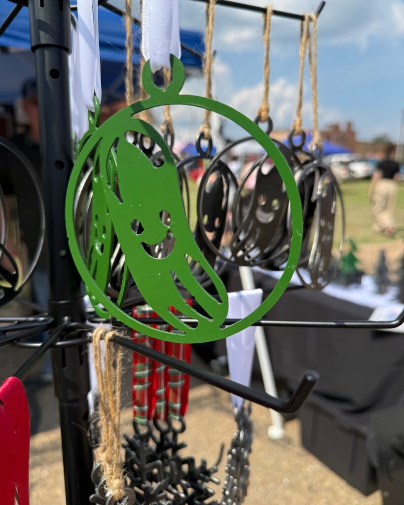 Green pickle-shaped metal ornament with a smiling face hanging on a vendor display rack at Texarkana Pickle Fest 2025 in Texarkana, Arkansas.