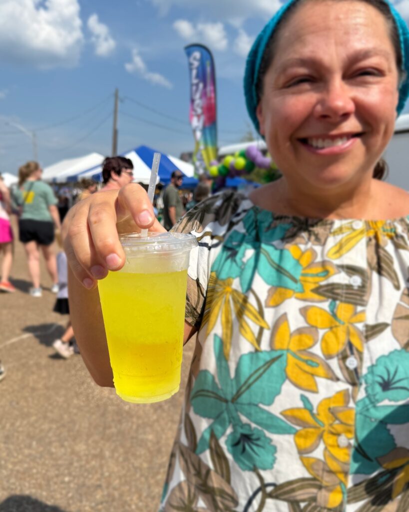 Smiling woman at Texarkana, Arkansas Pickle Fest 2025 holding a bright yellow pickle-flavored drink in a clear plastic cup with festival tents in the background.
