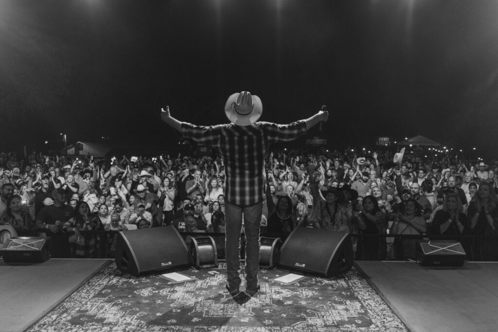 Black and white photo of country singer Tracy Lawrence standing on stage with arms raised toward a cheering crowd during a live outdoor concert at night.