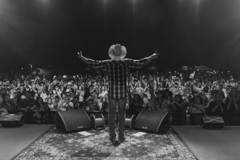 Black and white photo of country singer Tracy Lawrence standing on stage with arms raised toward a cheering crowd during a live outdoor concert at night.