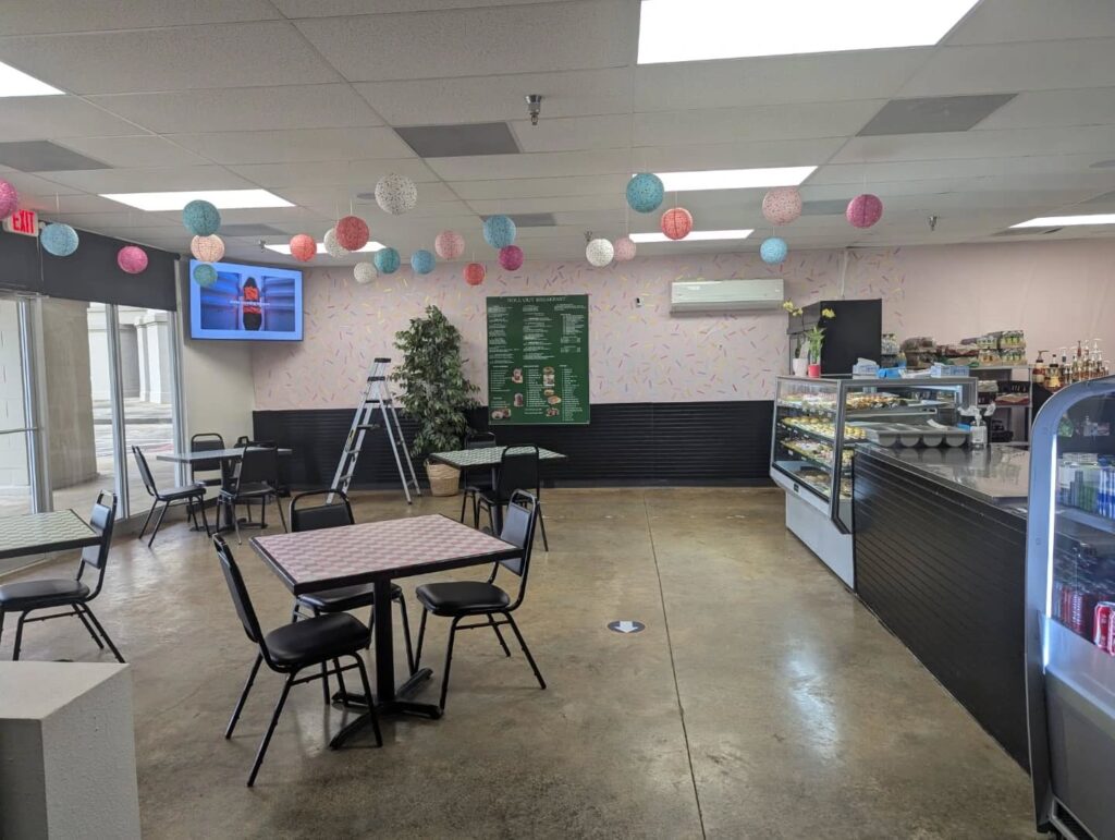 Inside Rolling Sweet Cafe, featuring pastel paper lanterns hanging from the ceiling, small tables with checkered tops, and a bright counter displaying pastries and desserts. The decor includes pink sprinkle-patterned walls, a green menu board, and a TV screen near the entrance, creating a cheerful and cozy atmosphere.