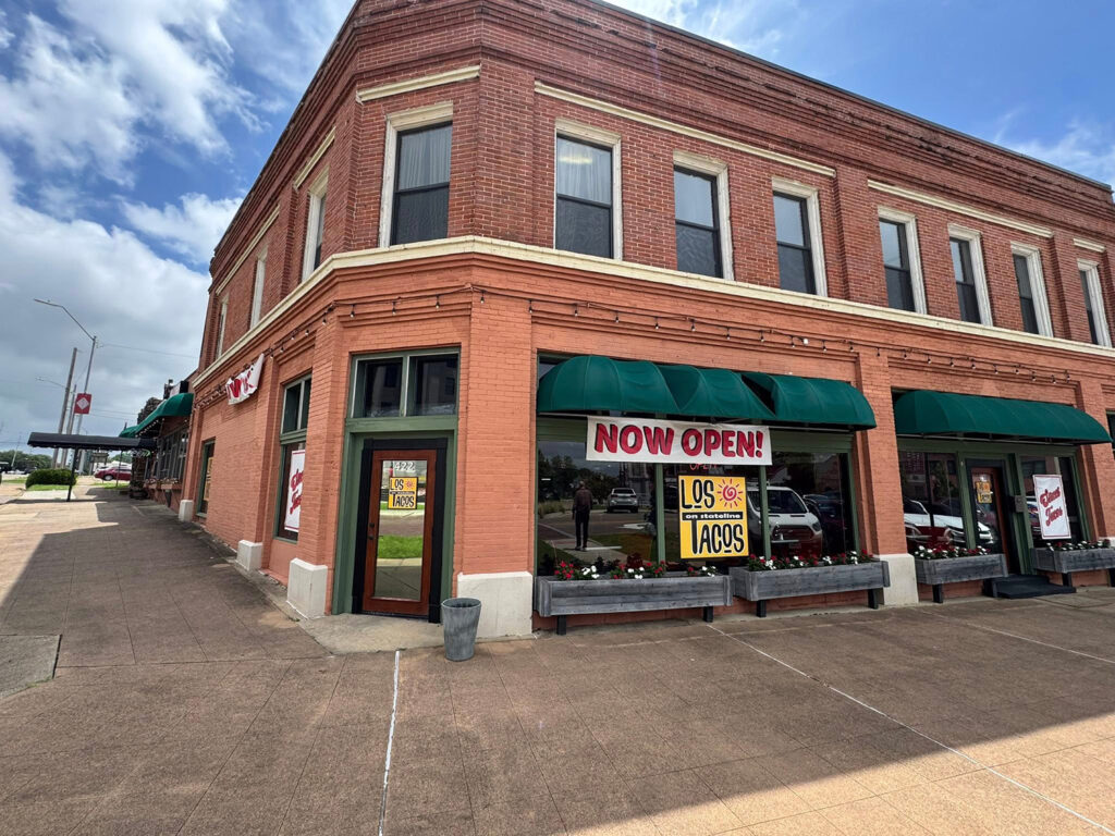 A brick corner building with green awnings and large windows displays a banner reading “NOW OPEN!” for Los Tacos on Stateline. The restaurant’s entrance and windows feature bright signage, and the sidewalk and blue sky create a welcoming downtown street view.