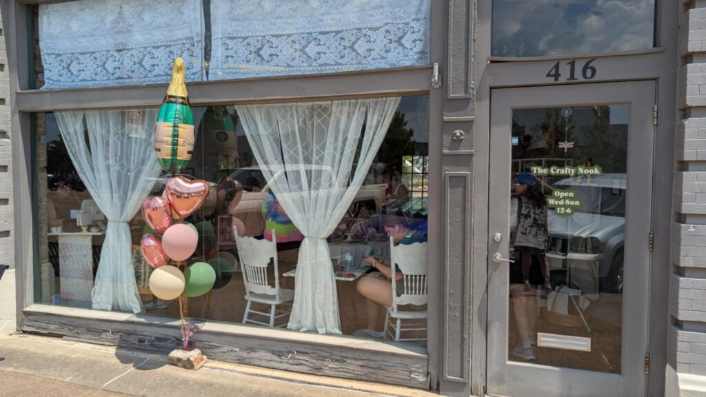 Street view of The Crafty Nook in Texarkana, Arkansas, showing the storefront with lace curtains, balloons, and people crafting inside through the front window at 416 N. State Line Avenue.