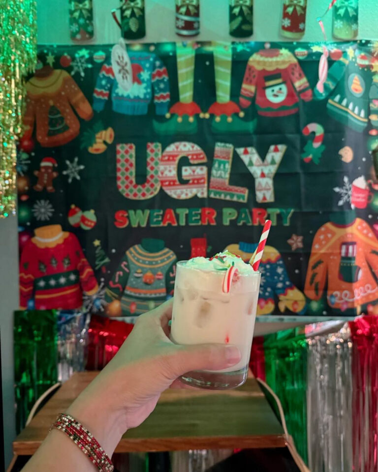 Hand holding a festive holiday cocktail with whipped cream and a candy cane straw in front of an “Ugly Sweater Party” backdrop decorated with colorful Christmas sweaters and tinsel at a Texarkana, Arkansas holiday pop-up.