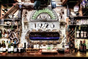 Vintage speakeasy bar interior with bottles, glassware, and industrial decor against a textured brick wall.