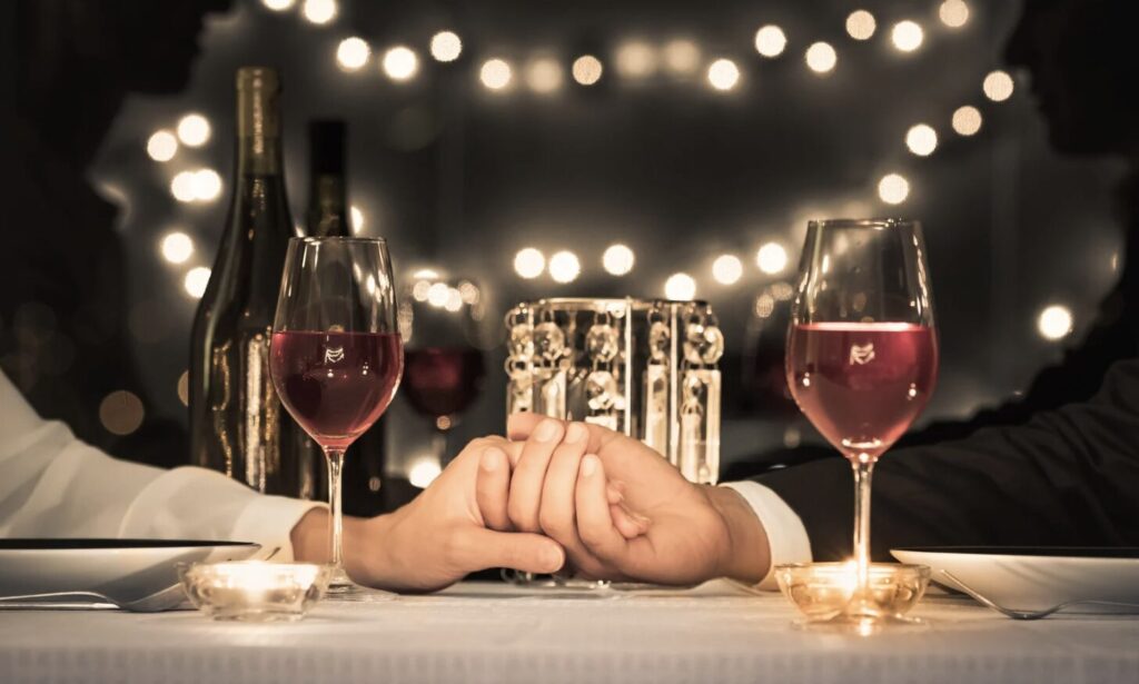Couple holding hands across a candlelit table with wine glasses and string lights.