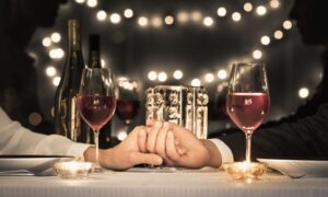 Couple holding hands across a candlelit table with wine glasses and string lights.